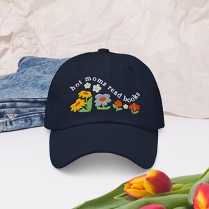 May include: A navy blue baseball cap with the text "hot moms read books" embroidered in white. The text is surrounded by colorful embroidered flowers.