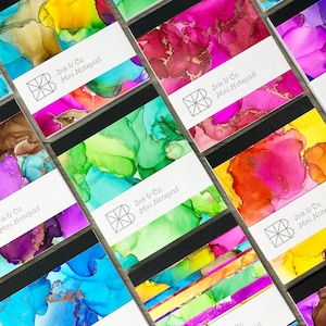 Puede incluir: Varios mini blocs de notas con diseños abstractos de acuarela en varios colores, incluyendo azul, verde, rosa y amarillo. Cada bloc de notas tiene un lomo negro y una etiqueta blanca con el texto "Ink & Co Mini Notepad" y un logotipo.