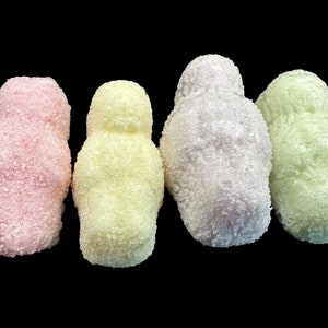 May include: Four sugar-coated marshmallow candies in the shape of people. The candies are pink, yellow, purple, and green.