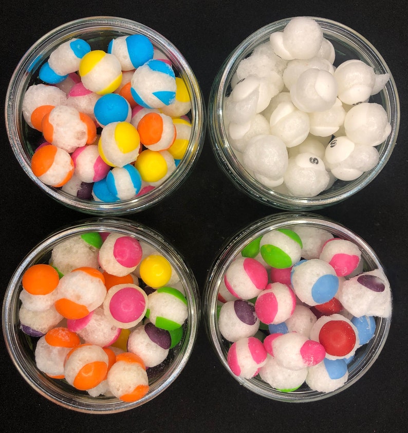 May include: Four glass jars filled with colorful candy. The jars are arranged in a square pattern on a black surface. The candy is a mix of white, red, blue, green, yellow, and orange.