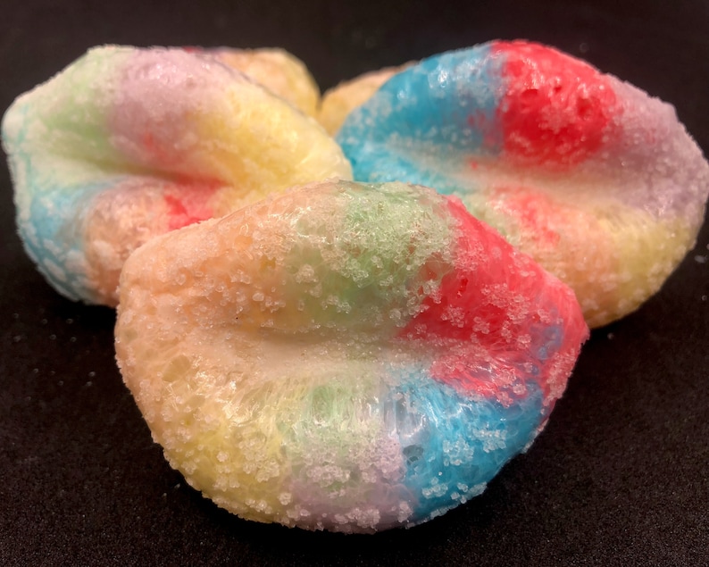 May include: Three colorful lip-shaped bath bombs with a sugar-like coating. The bath bombs are in shades of blue, pink, yellow, and green.