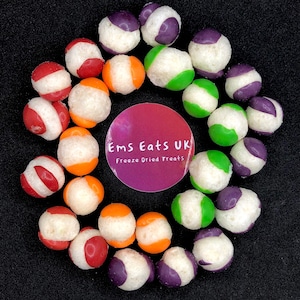 May include: A selection of freeze-dried fruit snacks arranged in a circle on a black background. The snacks are in various colours, including red, orange, green, and purple. The text "Ems Eats UK Freeze Dried Treats" is in the centre of the circle.