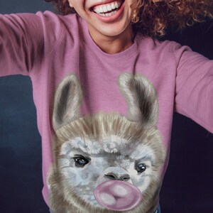 May include: A pink sweatshirt featuring a graphic of a llama wearing a pink bubble. The llama has a white face with brown fur around its eyes and ears.