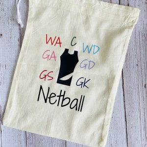 May include: A cream-coloured drawstring bag with the word "Netball" in black script. The bag features a black netball uniform graphic and colourful text: WA, GA, GS, C, WD, GD, and GK. A white drawstring is visible.