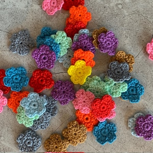May include: A selection of colourful, crocheted flower appliques in shades of pink, orange, red, yellow, blue, green, purple, grey, and brown. The flowers are scattered on a neutral surface.