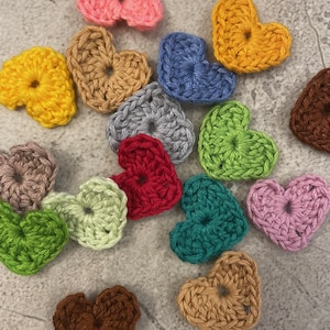 Könnte beinhalten: Eine Sammlung von 15 gehäkelten Herzen in verschiedenen Farben, darunter rosa, gelb, blau, grau, rot, grün, braun und lila.