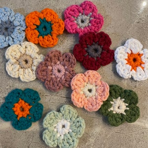 Może przedstawiać: Dziesięć kwiatów wykonanych na szydełku w różnych kolorach, w tym turkusowym, pomarańczowym, różowym, białym, czerwonym i zielonym. Kwiaty mają ten sam rozmiar i kształt, z pięcioma płatkami i kółkiem w środku.