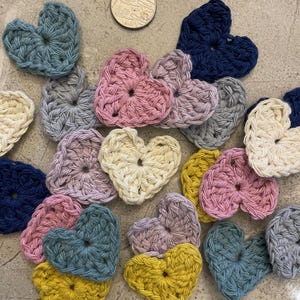 Puede incluir: Una colección de corazones tejidos a crochet en varios colores, incluyendo azul, rosa, amarillo, blanco y gris. Los corazones están dispuestos en un patrón aleatorio sobre una superficie gris.
