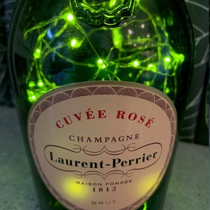 May include: A green glass bottle with a label that reads "Cuvée Rosé Champagne Laurent-Perrier Maison Fondée 1812 Brut". The bottle is decorated with white fairy lights.