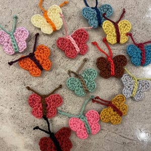 Puede incluir: Una colección de mariposas de ganchillo hechas a mano en varios tonos de rosa, amarillo, naranja, azul, marrón y verde. Cada mariposa tiene alas y antenas detalladas, perfectas para manualidades o decoración.