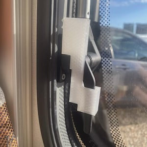 May include: A close-up of a window latch mechanism. The latch is white and attached to a black frame. The window has a dotted pattern and a view of the outside with a car visible.