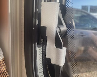 May include: A close-up of a window latch mechanism. The latch is white and attached to a black frame. The window has a dotted pattern and a view of the outside with a car visible.