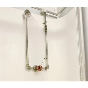 Op de afbeelding: Een zilveren douchekop met water dat naar beneden sproeit. De douchekop is verbonden met een reeks metalen buizen en fittingen, waaronder een klep met een rode en zwarte hendel. De douche is op een witte muur gemonteerd.