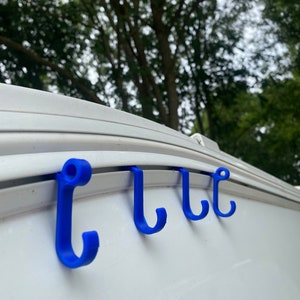 Op de afbeelding: Vier blauwe plastic haken zijn bevestigd aan een wit oppervlak. De haken zijn gelijkmatig verdeeld en hebben een gebogen ontwerp. De haken zijn waarschijnlijk bedoeld om items op te hangen. De achtergrond is wazig met groen gebladerte.