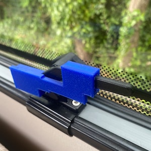 Pode incluir: Um objeto azul impresso em 3D, possivelmente um clipe ou suporte, fixado a uma barra preta. O objeto tem uma forma retangular com um braço saliente. O fundo mostra uma vista desfocada de folhagem verde.