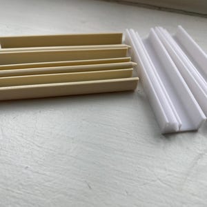 Könnte beinhalten: Eine Sammlung hellbeige und weißer Kunststoffleisten. Die beigefarbenen Teile sind gestapelt, während die weißen Teile in einer Reihe angeordnet sind. Diese architektonischen Elemente werden wahrscheinlich für dekorative oder funktionale Zwecke verwendet.