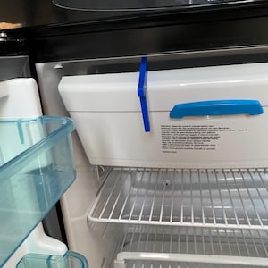 Può includere: Vista interna di un frigorifero con un contenitore bianco e dettagli blu. Il frigorifero ha ripiani in filo metallico e una parte superiore nera con il marchio "THETFORD". Il contenitore ha una maniglia blu e un fermo blu.