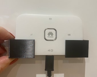Op de afbeelding: Een witte Huawei 4G mobiele hotspot met een zwarte oplaadkabel en twee zwarte rechthoekige houders. Het apparaat heeft een Huawei-logo en de tekst "4G". Het apparaat wordt door een hand vastgehouden.