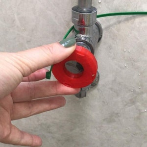 Op de afbeelding: Een rode kraanhendel met een groen touwtje eraan. De hendel wordt vastgehouden in de buurt van een chromen kraan op een lichtgekleurde muur. De hendel is rond met een gat in het midden.