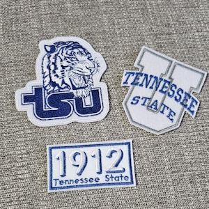 May include: Three embroidered patches featuring the Tennessee State University logo. The first patch shows a white tiger head with the letters "TSU" in blue. The second patch shows a white "U" with the words "Tennessee State" in blue. The third patch shows the year "1912" in white with the words "Tennessee State" in blue.