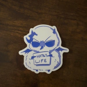 Puede incluir: Una pegatina azul y blanca de una criatura de dibujos animados con cuernos y alas que sostiene un cartel que dice "Hates Life".