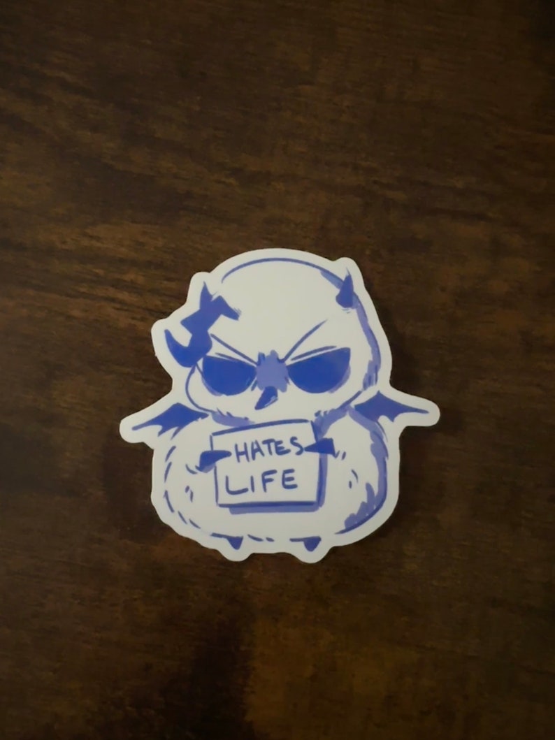 Puede incluir: Una pegatina azul y blanca de una criatura de dibujos animados con cuernos y alas que sostiene un cartel que dice "Hates Life".