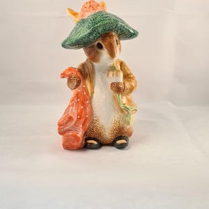 May include: A ceramic figurine of Peter Rabbit wearing a green hat and holding a red and white scarf. The figurine is brown and white and has a detailed design.