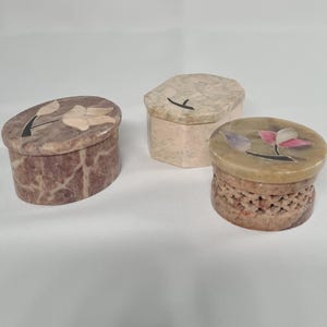 May include: Three small decorative boxes made of soapstone. The boxes are round and have different floral designs carved into the lids. The boxes are in various shades of brown, green, and white.