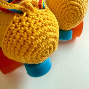 May include: A pair of yellow roller skates featuring bright yellow crocheted toe caps, multi-coloured striped laces in red, yellow, and blue, and light blue toe stops. The skates have a smooth yellow exterior finish and orange wheels.