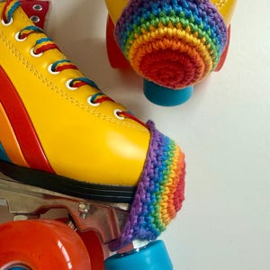 May include: A pair of yellow roller skates featuring rainbow-striped laces and a crocheted rainbow toe guard on each boot. The skates have bright orange wheels and light blue toe stops, with a vibrant multi-coloured design on the side panels.