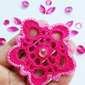 Puede incluir: Una flor de ganchillo rosa brillante hecha a mano con un borde rosa más claro y un centro de pedrería transparente. Alrededor de la flor hay varias piedras de estrás rosas en forma ovalada. La flor se sostiene sobre un fondo blanco.