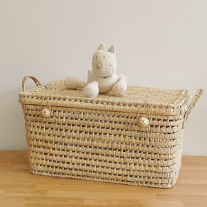 May include: A natural woven storage basket with a lid and two handles. The basket is made of light brown woven straw and has two small bells on the lid. A white stuffed animal is sitting on top of the lid.