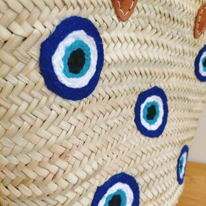 May include: A woven straw basket bag featuring multiple circular evil eye patches in shades of dark blue, light blue, white, and black. The bag includes tan leather handle attachments secured with visible white stitching on the upper rim.
