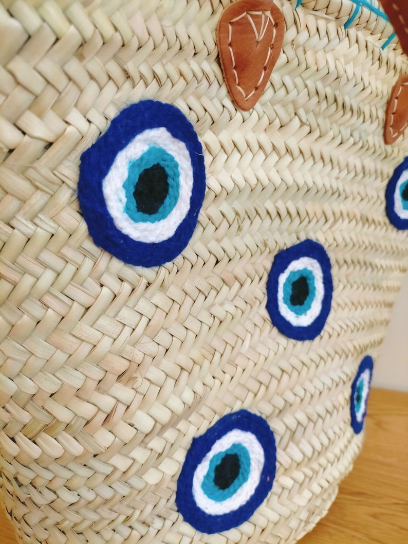 May include: A woven straw basket bag featuring multiple circular evil eye patches in shades of dark blue, light blue, white, and black. The bag includes tan leather handle attachments secured with visible white stitching on the upper rim.