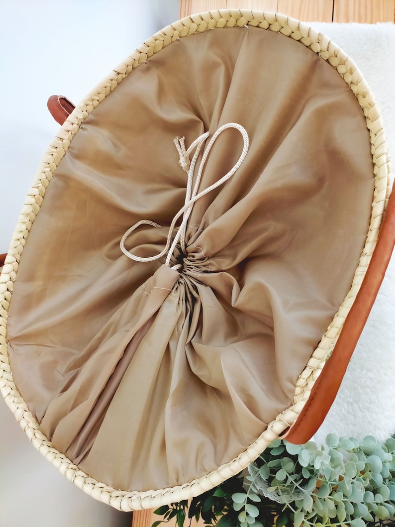 May include: A round, woven basket with a beige fabric liner and a drawstring closure. The basket is made of natural materials and has a light brown color.