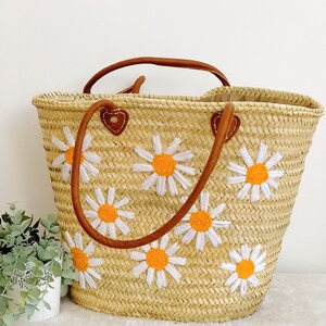 May include: A straw tote bag with brown leather handles and embroidered white daisies with orange centers.