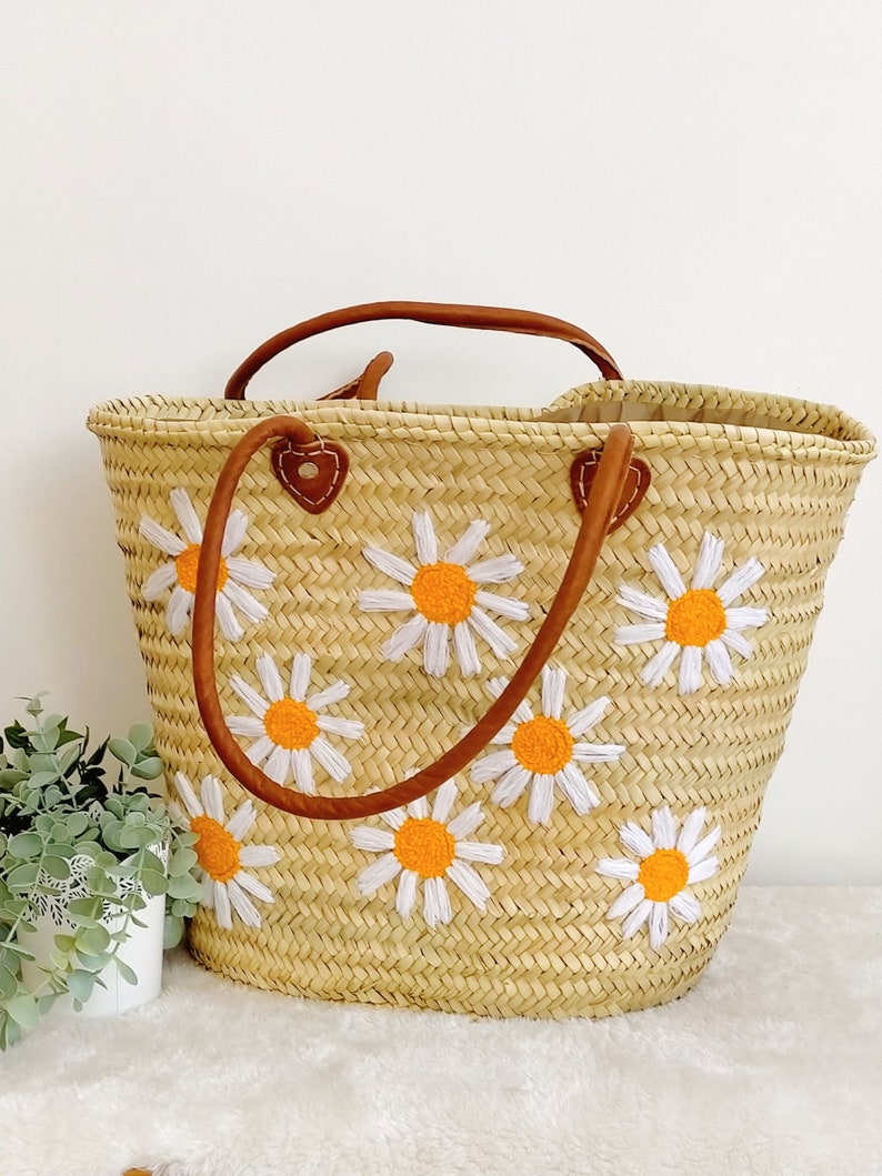 May include: A straw tote bag with brown leather handles and embroidered white daisies with orange centers.