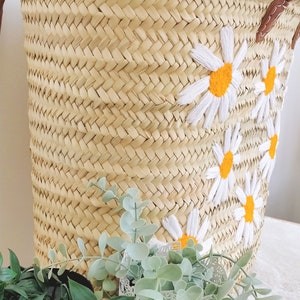 May include: A woven straw basket with a leather handle and white embroidered daisies with yellow centers. The basket is sitting on a table with green plants in the foreground.