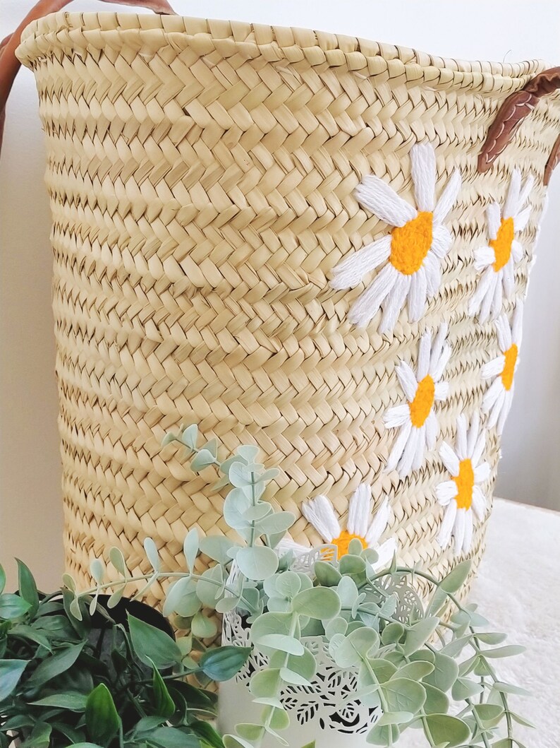 May include: A woven straw basket with a leather handle and white embroidered daisies with yellow centers. The basket is sitting on a table with green plants in the foreground.