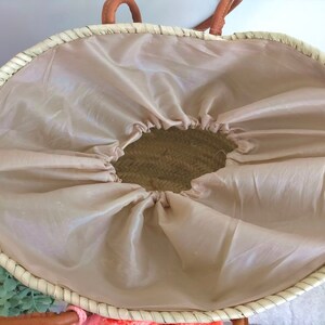 May include: A woven straw basket with a light beige fabric lining featuring a drawstring closure. The basket has a round opening and tan leather-coloured handles attached to the top rim. The interior of the basket is visible through the centre.