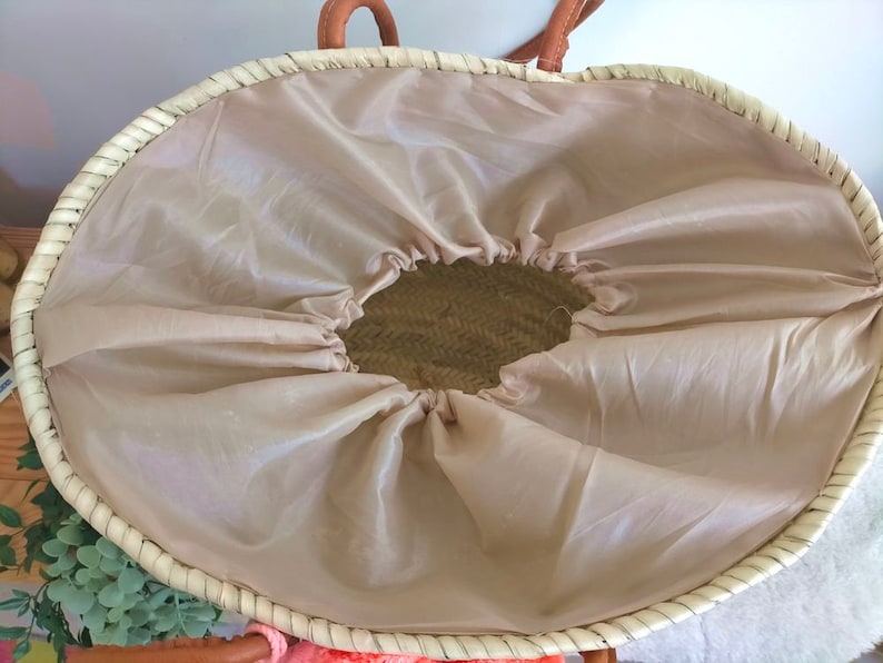 May include: A woven straw basket with a light beige fabric lining featuring a drawstring closure. The basket has a round opening and tan leather-coloured handles attached to the top rim. The interior of the basket is visible through the centre.