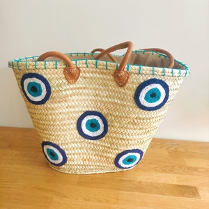 May include: A woven straw tote bag featuring four circular evil eye motifs in shades of dark blue, light blue, and white. The bag includes two brown leather handles attached with stitched leather patches and a teal stitched trim along the top edge.