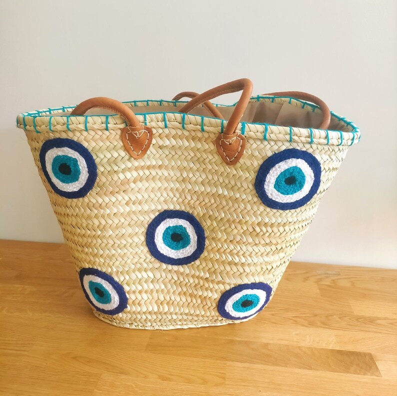 May include: A woven straw tote bag featuring four circular evil eye motifs in shades of dark blue, light blue, and white. The bag includes two brown leather handles attached with stitched leather patches and a teal stitched trim along the top edge.