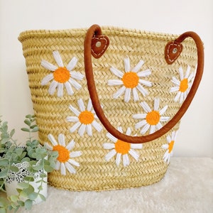 May include: A straw tote bag with brown leather handles and embroidered white daisies with orange centers.