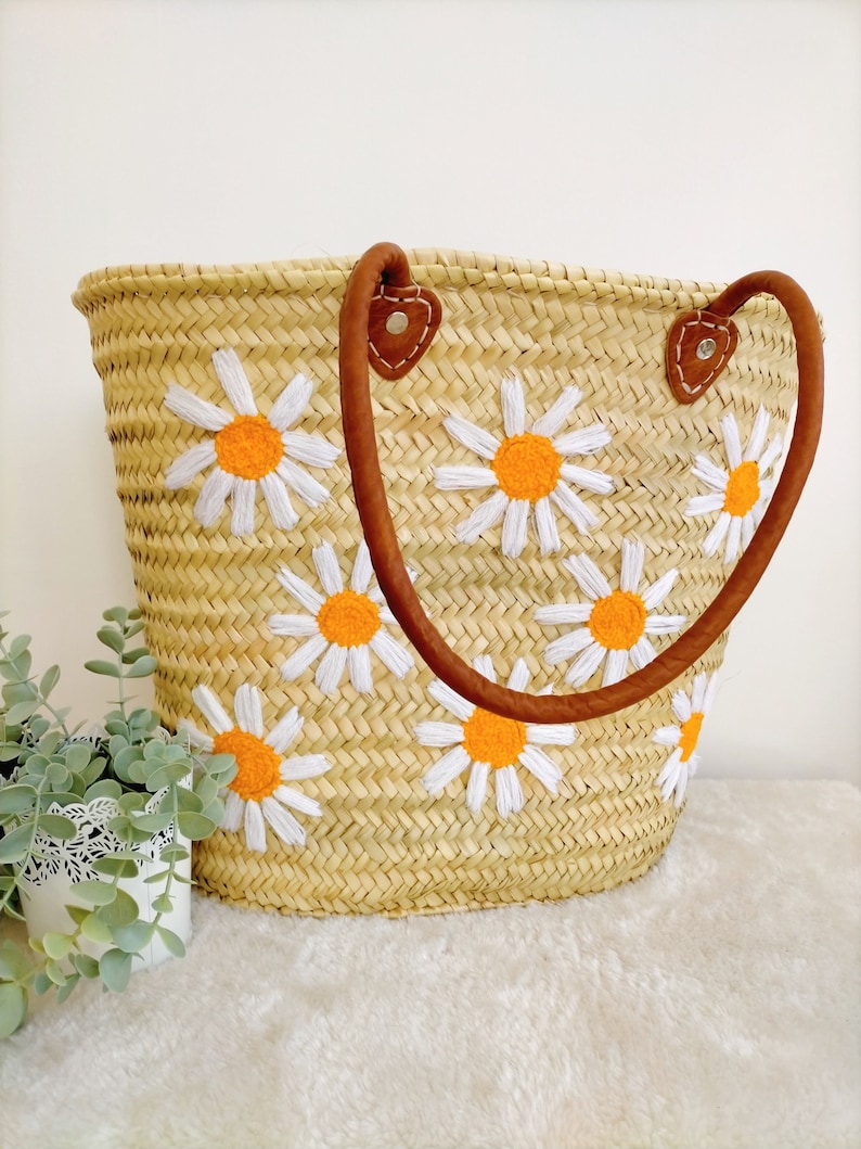 May include: A straw tote bag with brown leather handles and embroidered white daisies with orange centers.