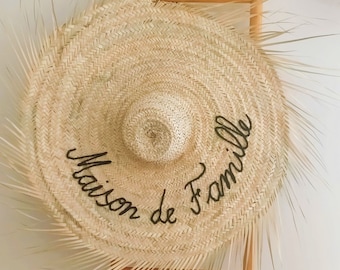 May include: A large, round, natural straw hat with a smaller, circular crown. The hat features the cursive text "Maison de Famille" in black. The hat is decorated with fringed edges and is displayed on a wooden ladder.