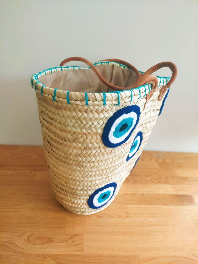 May include: A woven straw basket tote featuring three embroidered evil eye motifs in shades of navy blue, light blue, and white. The bag has a beige fabric lining, a teal stitched rim, and two brown leather handles attached to the top edge.
