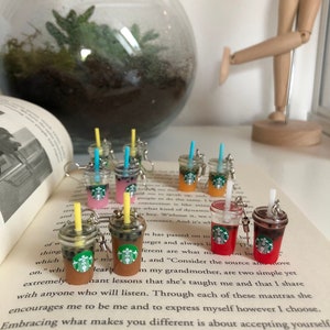 Könnte beinhalten: Miniatur-Anhänger im Starbucks-Stil mit verschiedenen Farben und Designs, darunter Braun, Rosa, Orange und Rot. Jeder Anhänger hat einen Strohhalm und ein Starbucks-Logo, an einem Schlüsselanhänger befestigt.