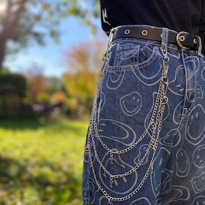 Può includere: Jeans blu con un motivo di faccine bianche. I jeans sono abbinati a una cintura nera e una catena argentata con ciondoli. Lo sfondo è una scena all'aperto sfocata.