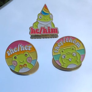 Könnte beinhalten: Drei Frosch-Emaille-Pins mit Regenbogenstreifen und verschiedenen Pronomen-Etiketten: sie/ihr, er/ihm und sie/ihnen. Jeder Pin zeigt einen Cartoon-Frosch mit einem anderen Ausdruck.
